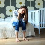 Young tired woman sitting on the bed near children’s cot.